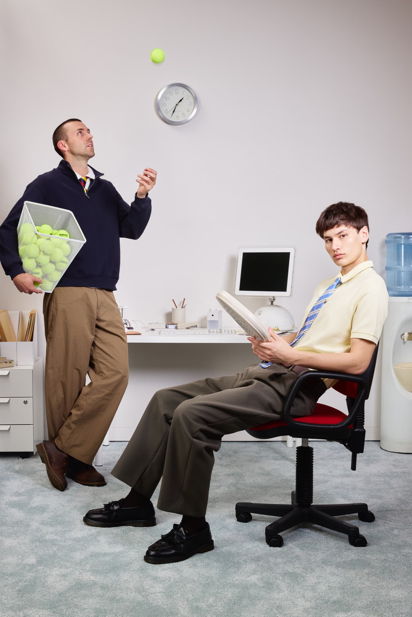 Two colleagues in a retro office, one tossing a ball while the other holds a padel racket
